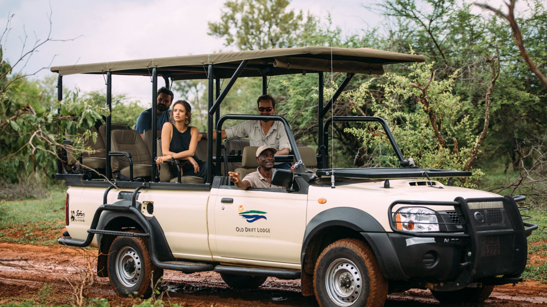 Game drive safari vehicle with a canopy carries four passengers on a red dirt road.