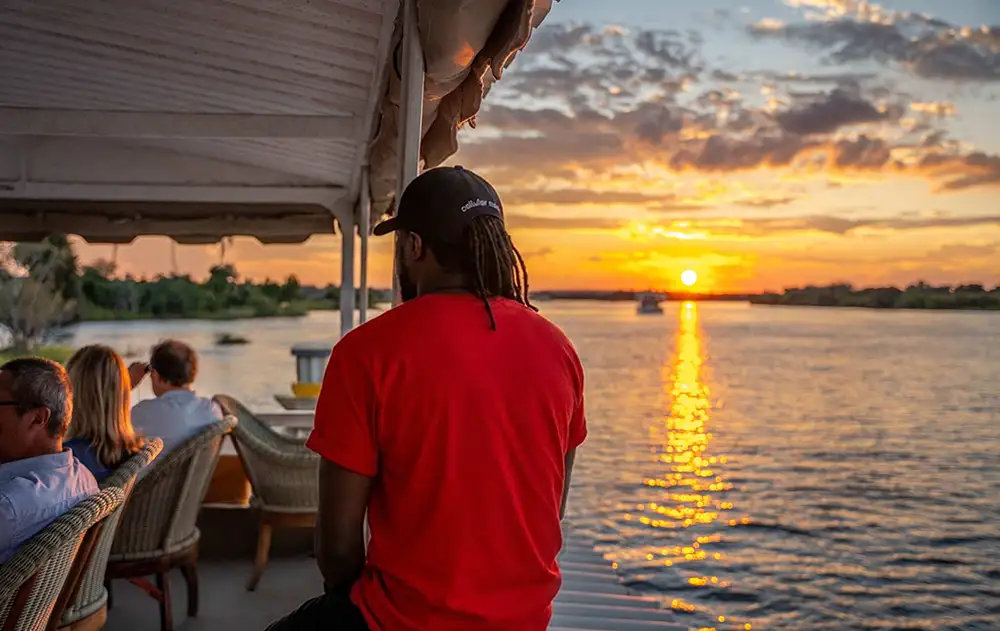 Open deck of the cruise boat at golden hour — wide river, sunset sky, silhouettes of guests or wildlife on the bank. Warm tones. Avoid tight portrait crops here — the landscape is the point.