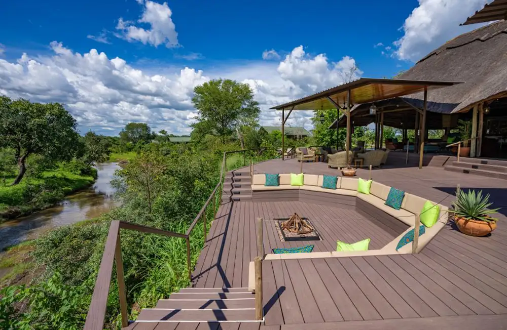 Wide shot of the lodge deck and pool with Masuwe River backdrop - green riverine trees, calm water, preferably morning light. No people needed - let the setting speak.