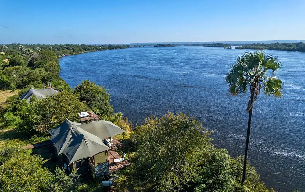 Establishing shot of Old Drift Lodge from the river or riverbank — canvas suites in the trees, Zambezi in the foreground, national park bush. Morning or late afternoon.