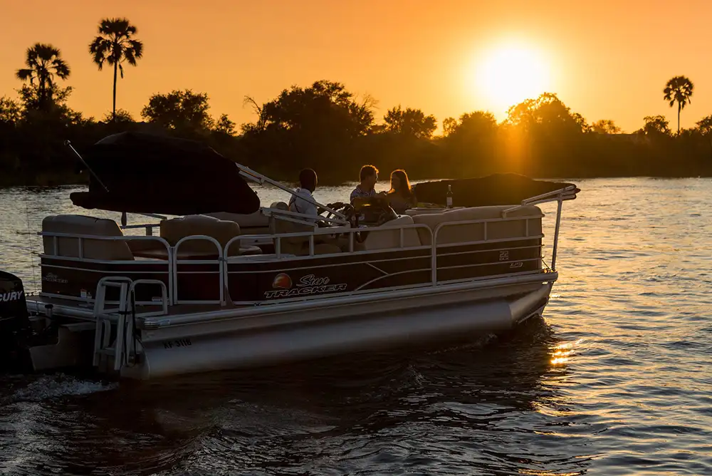 Pontoon boat on the Zambezi with elephant or other large wildlife on the bank — wide shot, warm late-afternoon light, national park bush in background.
