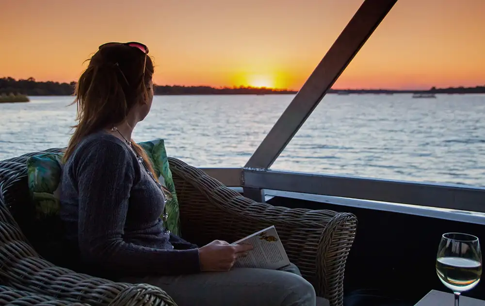 Upper deck of the Malachite at golden hour — wide river, sunset sky, silhouettes of guests or wildlife on the bank. Warm tones. The boat and the landscape together.