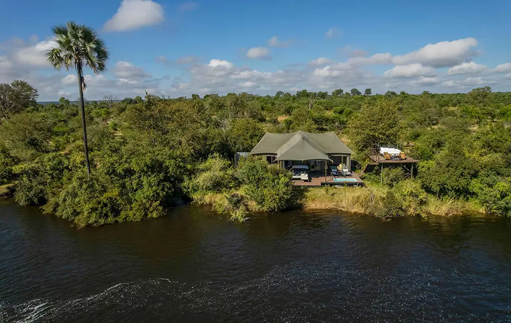 Establishing shot of Old Drift Lodge from the riverbank or water — suites in the trees, Zambezi in the foreground, national park bush. Morning or late afternoon.