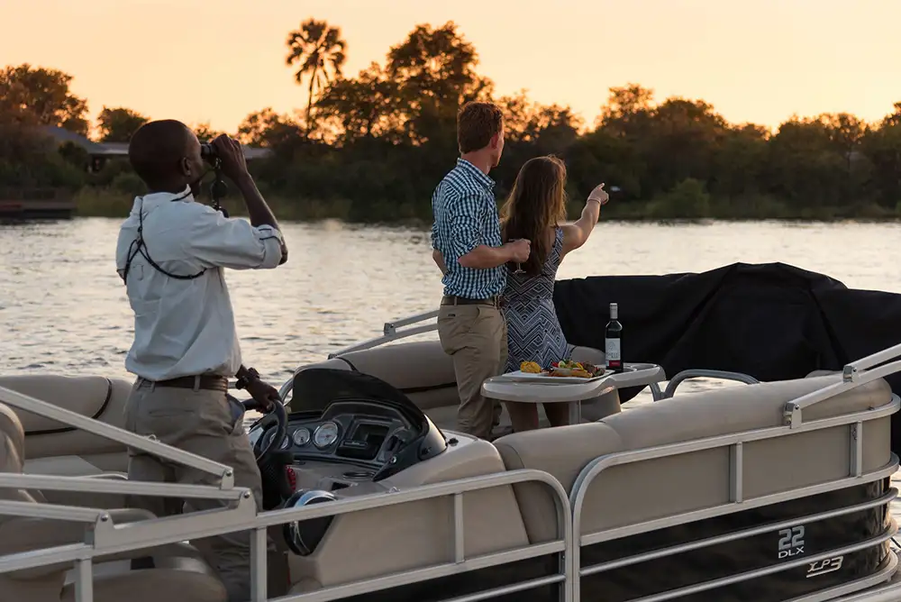 boat on the Zambezi with elephant or buffalo at the water's edge — wide shot, warm light, national park bush in background. Wildlife should be the subject, not the guests.